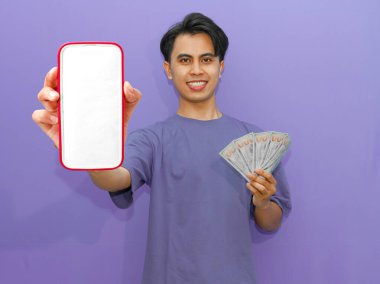Gülümseyen Asyalı genç bir adam, canlı mor bir arkaplan üzerine kurulmuş banknotları tutarken boş bir cep telefonu ekranı gösteriyor..
