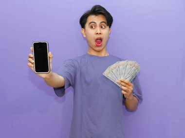 Şok olmuş bir ifadeyle genç bir Asyalı elinde boş bir cep telefonu ekranı ve parlak mor bir arka plana karşı banknotlar tutuyor..