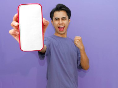 Heyecanlı Asyalı genç adam başarıyı kutluyor, boş bir cep telefonu ekranı gösteriyor, canlı mor bir arka planda yumruklarını sıkarak taklit ediyor..