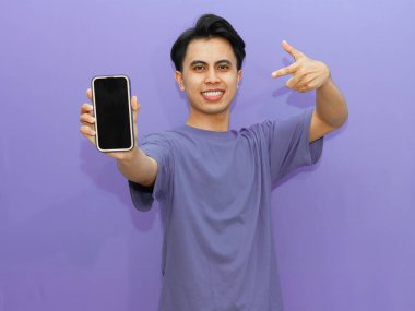 Heyecanlı genç Asyalı adam canlı mor bir arka planda boş bir cep telefonu ekranını işaret ediyor. Reklam için mükemmel..