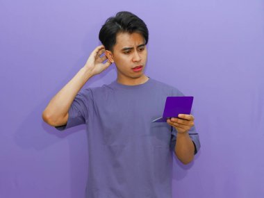 Zavallı Asyalı genç adam, kafasını mor bir arka plana sürterek tasarruf defterini tutarken endişeli görünüyor..