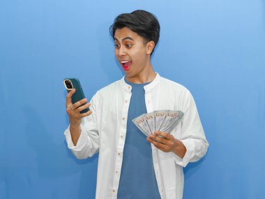 Elinde banknot tutarken cep telefonu kullanan, mavi arka planda oturan, komik bir Asyalı genç adam..