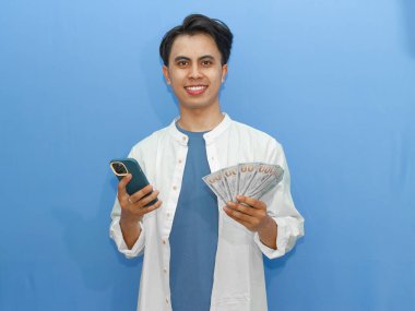 Kendine güvenen genç Asyalı bir adam elinde cep telefonu ve banknotlarla gülümsüyor. Mavi arka planda reklam yapmak için mükemmel..