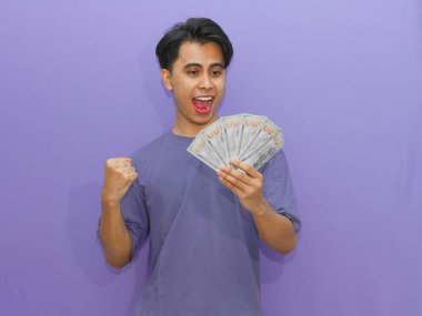 Heyecanlı genç Asyalı adam elinde para tutuyor, neşeli bir ifadeyle başarıyı kutluyor, canlı mor bir arka planda reklam ve promosyonlar için mükemmel..