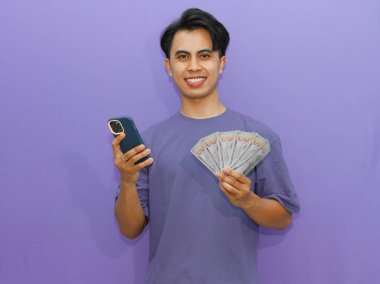 Genç Asyalı adam, elinde mor bir fon, reklam, finans ve başarı temaları için mükemmel bir para ve telefon tutarken güvenle gülümsüyor..