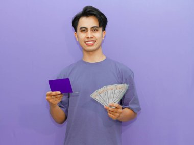 Asyalı genç adam elinde para ve tasarruf defteri tutarken banka tasarruflarına bakarken mutlu bir ifade sergiliyor. Finans ve başarı temaları için mükemmel.