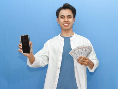 Sıradan mavi tişörtlü genç bir Asyalı kendine güvenen boş bir cep telefonu ve banknotları tutuyor, finansal reklamlar için ideal..
