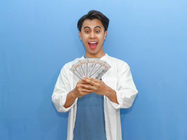 Mavi gömlekli genç Asyalı adam elinde heyecanlı bir ifadeyle banknotlar tutarak finansal başarıyı kutluyor. Zenginlik ve refah kavramları üzerine reklamlar için ideal..