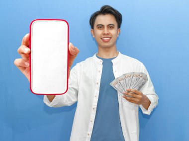 Sıradan mavi tişörtlü genç bir Asyalı kendine güvenen boş bir cep telefonu ve banknotları tutuyor, finansal reklamlar için ideal..