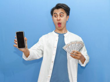 Sıradan mavi gömlekli genç bir Asyalı elinde banknotlar tutarken boş bir cep telefonu ekranı gösteriyor ve şaşırdığını gösteriyor. Finansal reklamlar için ideal.