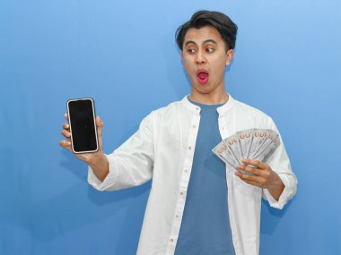 Sıradan mavi tişörtlü genç bir Asyalı elinde boş bir telefon ve banknotla komik bir şok ifadesi sergiliyor. Finansal reklamlar için ideal..