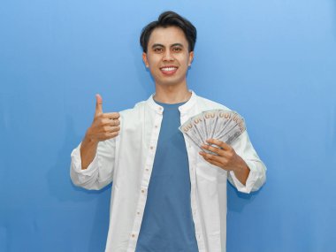 Mavi gömlekli genç Asyalı bir adam nakit para tutuyor ve kendine güvenen bir gülümsemeyle onaylıyor. Finansal reklamcılık ve tanıtım kavramları için ideal..