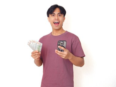 Mutlu Asyalı genç adam elinde akıllı telefon ve nakit tutarak şaşırdı. Heyecanlı ifade tarzı, sade bir arka planda izole, finans, iş ve reklam teması için mükemmel..