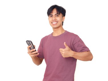 Gülümseyen Asyalı genç adam akıllı telefonu tutuyor ve baş parmak işareti yapıyor, beyaz arka planda izole edilmiş, başarı kavramı, onay, teknoloji ve iletişim..