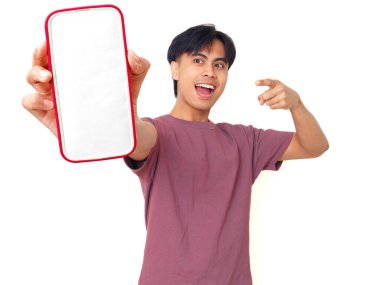 Mutlu Asyalı genç adam gülümsüyor ve boş akıllı telefon ekranına işaret ediyor. İzole edilmiş beyaz arka planda reklam için kopya alanı sunuyor..