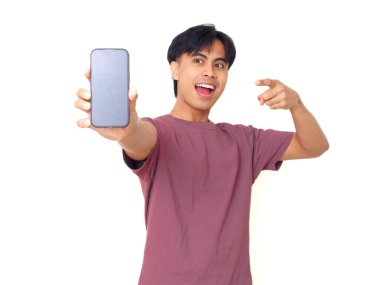 Mutlu Asyalı genç adam gülümsüyor ve boş akıllı telefon ekranına işaret ediyor. İzole edilmiş beyaz arka planda reklam için kopya alanı sunuyor..
