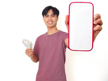 Heyecanlı Asyalı genç adam elinde nakit para tutuyor ve beyaz arka planda finansal uygulama ya da reklam için fotokopi alanı olan boş akıllı telefon ekranı gösteriyor..