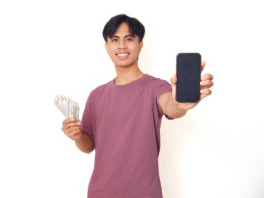Heyecanlı Asyalı genç adam elinde nakit para tutuyor ve beyaz arka planda finansal uygulama ya da reklam için fotokopi alanı olan boş akıllı telefon ekranı gösteriyor..
