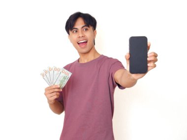 Heyecanlı Asyalı genç adam elinde nakit para tutuyor ve beyaz arka planda finansal uygulama ya da reklam için fotokopi alanı olan boş akıllı telefon ekranı gösteriyor..