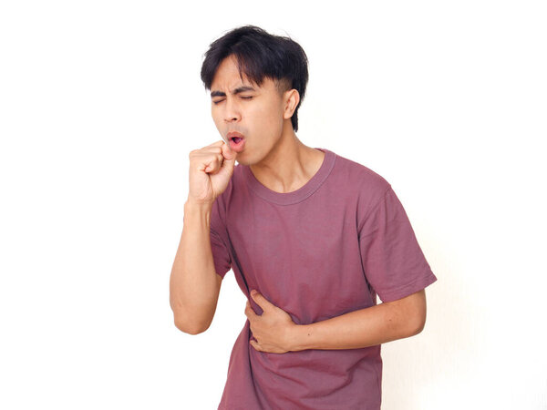 Young Asian man coughing and holding stomach with painful expression on plain background, suitable for health, illness, medical concept, and healthcare advertising.