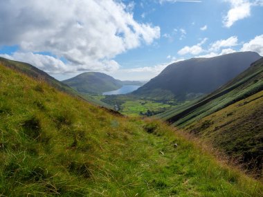 Panoramik bir manzara, bulutlu bir gökyüzünün altındaki manzarayı çerçeveleyen görkemli dağlarla, dingin bir göle uzanan yuvarlanan yeşil tepeleri yakalar..