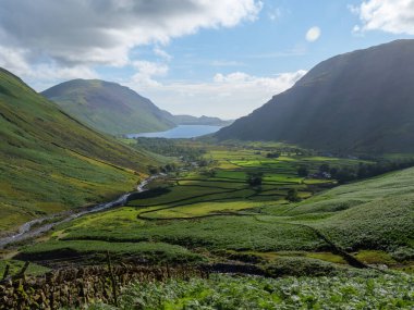 Parlak güneş ışığı ve dağınık bulutlarla aydınlatılmış, arka planda dingin bir göl bulunan yeşil tepeler ve vadileri saran çarpıcı bir manzara sergiliyor..