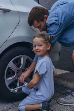 Sevimli küçük kız sıcak bir günde babasının aile arabasının tekerleğini değiştirmesine yardım ediyor. Yüksek kalite fotoğraf