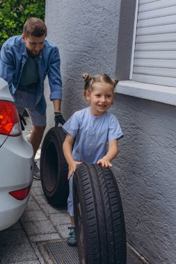 Sevimli küçük kız sıcak bir günde babasının aile arabasının tekerleğini değiştirmesine yardım ediyor. Yüksek kalite fotoğraf