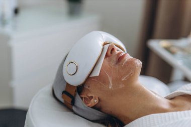 Genç bir kadın, sağlık hizmetlerini ön plana çıkaran bir cihazla göz masajı yaptırıyor. Yüksek kalite fotoğraf