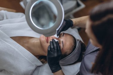 Plastik eldivenli kozmetik uzmanı LED ışıklı lupa ile yüz temizleme ameliyatı yapıyor. Yüksek kalite fotoğraf