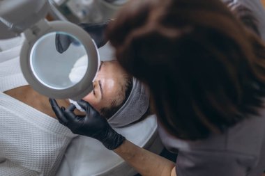 Plastik eldivenli kozmetolog, LED ışıklı lupa ile büyüteç kullanarak yüz temizliği yapıyor. Yüksek kalite fotoğraf