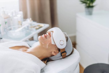 Genç bir kadın, sağlık hizmetlerini ön plana çıkaran bir cihazla göz masajı yaptırıyor. Yüksek kalite fotoğraf