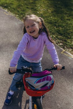  5 yaşındaki bir kız, parkta bisikletini sürüyor, gülümsüyor ve güneşli, yeşil ortamın tadını çıkarıyor. Yüksek kalite fotoğraf