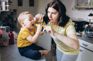 Anne oğlunu mutfakta kaşıkla besliyor, neşeli ve sevgi dolu bir annelik anını paylaşıyor. Yüksek kalite fotoğraf