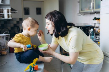 Anne oğlunu mutfakta kaşıkla besliyor, neşeli ve sevgi dolu bir annelik anını paylaşıyor. Yüksek kalite fotoğraf