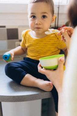 Anne oğlunu mutfakta kaşıkla besliyor, neşeli ve sevgi dolu bir annelik anını paylaşıyor. Yüksek kalite fotoğraf