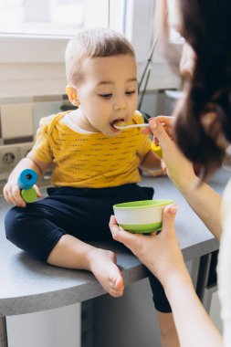 Anne oğlunu mutfakta kaşıkla besliyor, neşeli ve sevgi dolu bir annelik anını paylaşıyor. Yüksek kalite fotoğraf
