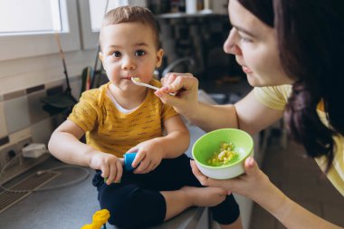 Anne oğlunu mutfakta kaşıkla besliyor, neşeli ve sevgi dolu bir annelik anını paylaşıyor. Yüksek kalite fotoğraf