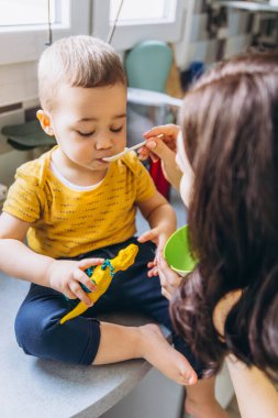Anne oğlunu mutfakta kaşıkla besliyor, neşeli ve sevgi dolu bir annelik anını paylaşıyor. Yüksek kalite fotoğraf