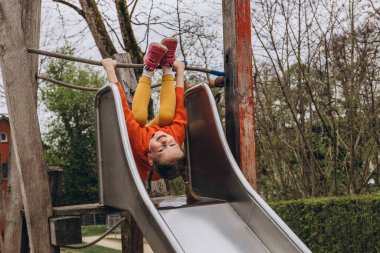 Çocuk büyük bir gülümsemeyle oyun parkından kayıyor, dışarıda neşeli ve kaygısız bir anın tadını çıkarıyor. Yüksek kalite fotoğraf