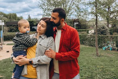 Genç bir aile parkın dışında birlikte vakit geçiriyor. Anne kucağında bir bebekle dolaşırken, baba onları kucaklıyor, günlük giysiler içinde, doğada huzurlu bir günün tadını çıkarıyor. Yüksek kalite fotoğraf