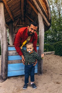 Baba ve küçük çocuk açık hava oyun parkında tahta bir oyun evinde birlikte oynuyorlar. Çocuk, doğal ahşap dokular ve yeşilliklerle çevrili sarı bir kepçeden kum döküyor. Yüksek kalite fotoğraf