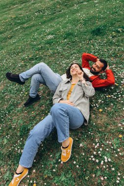 İki insan, kadın ve erkek yazın çimenlikte küçük kır çiçekleriyle birlikte dinleniyor, dışarıda huzurlu bir anın tadını çıkarıyorlar. Günlük giysiler ve doğal çevre sakin ve otantik bir ortam yaratıyor. Yüksek kalite fotoğraf