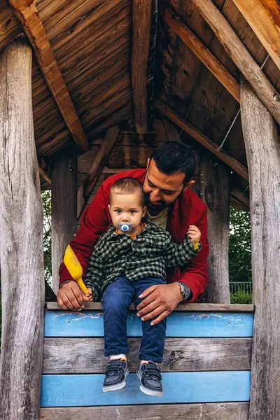 Baba ve küçük çocuk açık hava oyun parkında tahta bir oyun evinde birlikte oynuyorlar. Çocuk, doğal ahşap dokular ve yeşilliklerle çevrili sarı bir kepçeden kum döküyor. Yüksek kalite fotoğraf