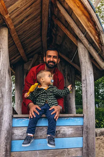 Baba ve küçük çocuk açık hava oyun parkında tahta bir oyun evinde birlikte oynuyorlar. Çocuk, doğal ahşap dokular ve yeşilliklerle çevrili sarı bir kepçeden kum döküyor. Yüksek kalite fotoğraf
