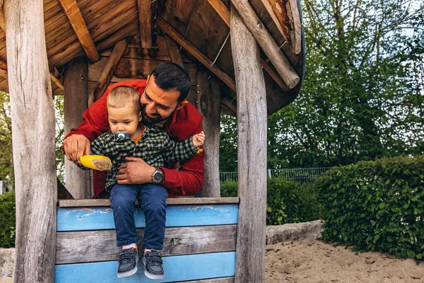 Baba ve küçük çocuk açık hava oyun parkında tahta bir oyun evinde birlikte oynuyorlar. Çocuk, doğal ahşap dokular ve yeşilliklerle çevrili sarı bir kepçeden kum döküyor. Yüksek kalite fotoğraf
