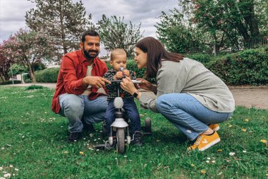 Parkta iki yaşındaki oğulları olan bir anne ve baba. Bebek küçük bir bisiklete biniyor, dışarıda eğleniyor, eğleniyor ve öğreniyor. Yüksek kalite fotoğraf