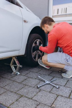Evinin önünde lastik değiştiren bir adamın yakın çekimi. Araç kriko ile kaldırılır, aletler yakınlarda durur. Olay yeri, gündüz vakti elle tamir edilen bir anı yakalıyor. Yüksek kalite fotoğraf