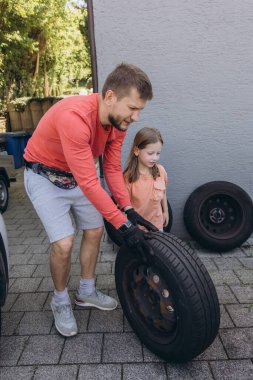 Evinin önünde lastik değiştiren bir adamın yakın çekimi. Araç kriko ile kaldırılır, aletler yakınlarda durur. Olay yeri, gündüz vakti elle tamir edilen bir anı yakalıyor. Yüksek kalite fotoğraf