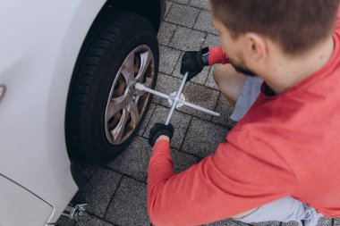 Evinin önünde lastik değiştiren bir adamın yakın çekimi. Araç kriko ile kaldırılır, aletler yakınlarda durur. Olay yeri, gündüz vakti elle tamir edilen bir anı yakalıyor. Yüksek kalite fotoğraf